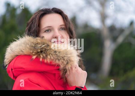 Junge Frau versteckt ihr Gesicht vor Kälte in Kapuze der roten Winterjacke. Porträt der Frau vor dem Hintergrund von immergrünen Nadelwald. Stockfoto