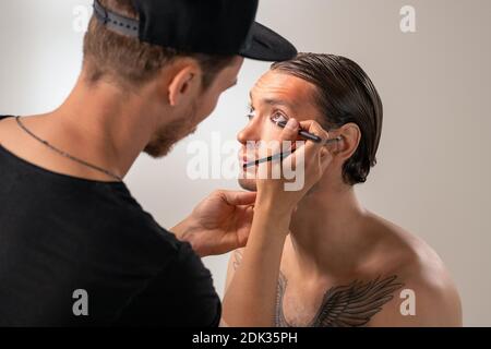 Über der Schulter Ansicht von MUA Künstler in Kappe Anwendung Eyeliner An jungen bärtigen Schauspieler mit Tätowierung auf Brust im Studio Stockfoto
