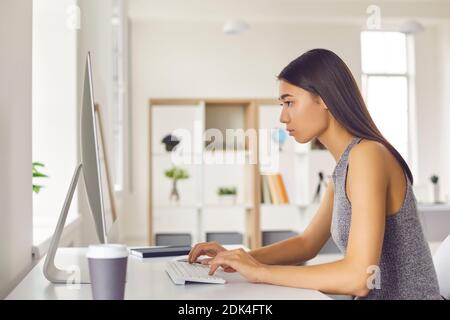 Junge hübsche asiatische Frau Büroangestellte sitzen am Computer und Monitor und arbeiten online, Seitenansicht Stockfoto