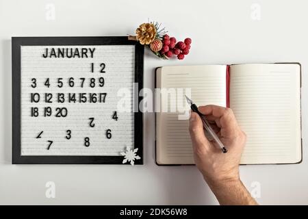 Männliche Hand mit einem Stift und einem Notizbuch für Notizen von Zielen und Plänen für das neue Jahr, Kalender und Weihnachtsbaumschmuck auf dem Desktop Stockfoto