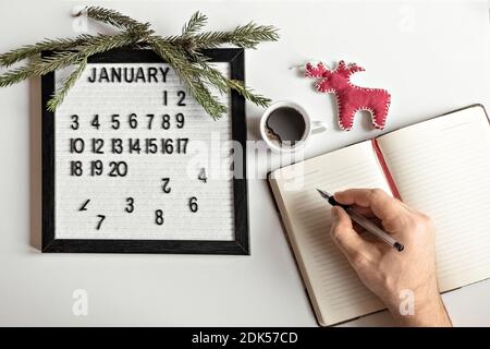 Männliche Hand mit einem Stift und einem Notizbuch für Notizen von Zielen und Plänen für das neue Jahr, Kalender und Weihnachtsbaumschmuck auf dem Desktop Stockfoto