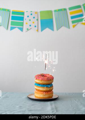 Geburtstags Donuts Kuchen mit bengalen Licht oder Sparkler. Stapel von leckeren Donuts über Tisch mit Party-Flagge Girlande auf Hintergrund. Vertikal. Speicherplatz kopieren Stockfoto