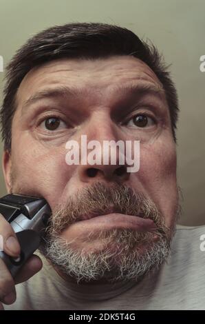 Ein erwachsener Mann rasiert sich mit einem elektrischen Rasiermesser. Ungewöhnliches Porträt eines bärtigen Mannes mit grauem Haar. Grimassen vor der Kamera Stockfoto