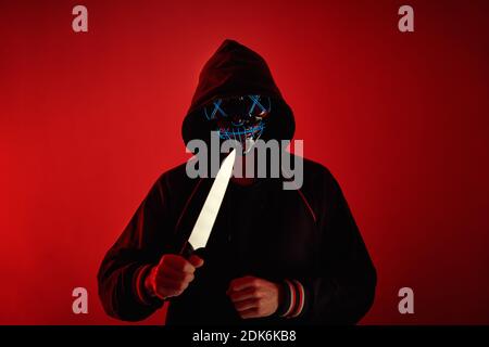 Mann im Hoodie mit gruseliger Maske im Gesicht hält Messer in der Hand auf rotem Hintergrund. Kriminelles Konzept Stockfoto