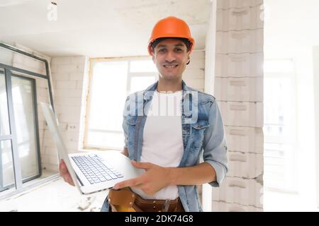 Schöner Architekt oder Vorarbeiter im Helm, der mit Laptop arbeitet Die Wohnung Zeichnungen an der Struktur weiß Interieur Stockfoto