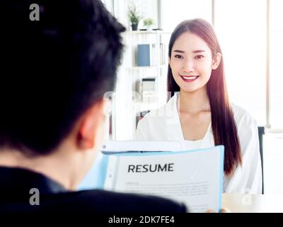 Junge attraktive asiatische Frau während Job-Interview mit Geschäftsmann in Anzug Lesung Lebenslauf im Büro. Schöne weibliche Kandidatin lächelt während intervie Stockfoto
