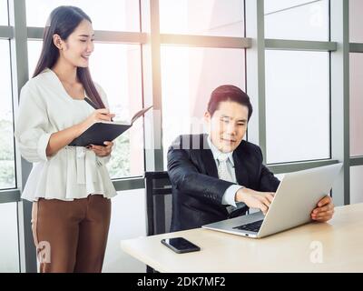 Asiatischer Geschäftsmann, Manager in Anzug zeigt auf Bildschirm des Laptop-Computer und junge lächelnde Frau Sekretärin, die Notiz nimmt, arbeiten zusammen für den Erfolg. Stockfoto