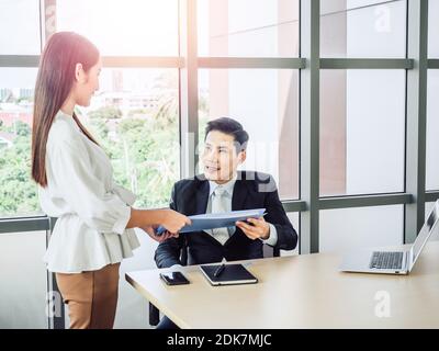 Asiatischer Geschäftsmann, Manager im Anzug, das Dokument von der jungen schönen Frau Sekretärin am Schreibtisch in der Nähe des Glasfensters, die für s Stockfoto