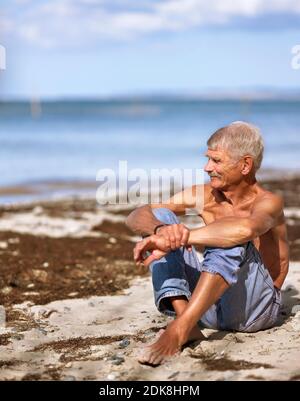 Älterer Mann in Jeans sitzt am Sandstrand Stockfoto