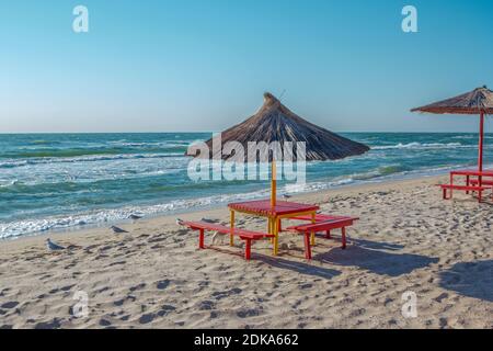 Sommerlandschaft: Tisch, Bänke und Sonnenschirm am Strand. Goldener Sand, stürmisches und winkendes Meer und blauer Himmel. Stockfoto