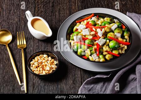 Makkaroni-Salat mit Brokkoli-Sprossen, Paprika-Streifen, Feta und Erdnuss in einer schwarzen Schüssel mit Erdnusssauce auf einem Holztisch, horizontale Ansicht von ab Stockfoto