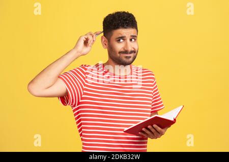 Verärgert nachdenklichen Mann in gestreiften T-Shirt reiben Hinterkopf mit Bleistift halten Notebook in der Hand, schriftlich Pläne für die Zukunft, machen, um Liste zu tun. ICH Stockfoto