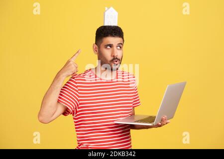 Lustiger bärtiger Mann in gestreiftem T-Shirt, der auf das Papierhaus auf dem Kopf zeigt und einen Laptop hält, von zu Hause aus arbeitet, freiberuflich arbeitet, Telearbeit. Indoo Stockfoto