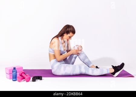 In voller Länge Seitenansicht verärgert athletische Frau in weißer Sportkleidung halten Knie auf Gummimatte sitzen, Gefühl Schmerzen, verletztes Bein während des Trainings. Innen s Stockfoto