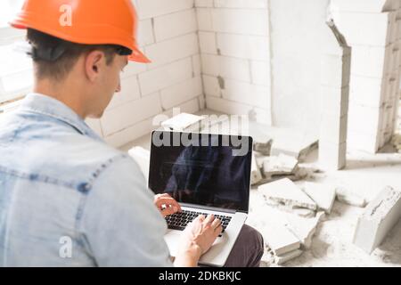 Schöner Architekt oder Vorarbeiter im Helm, der mit Laptop arbeitet Die Wohnung Zeichnungen an der Struktur weiß Interieur Stockfoto