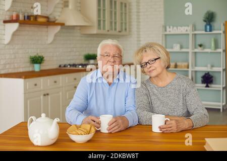 Seniorenpaar sitzt in der Küche und schaut auf die Kamera und trinkt Tee oder Kaffee mit Keksen. Stockfoto