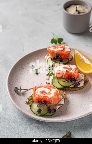 Mini-Sandwiches mit geräuchertem Lachs, Frischkäse, Gurke und Microgreen auf Roggenbrot, grauer Steinhintergrund. Vorspeise mit Lachs und Käse c Stockfoto