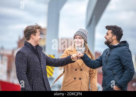 Two men and a woman greeting at Corona time, without everyday masks, out and about in the city in the cold season, Stock Photo