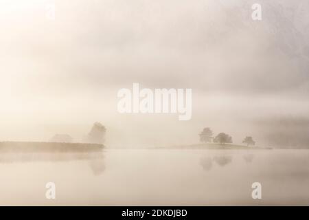 Herbststimmung an einem kleinen See in den bayerischen Alpen, im Hintergrund das Karwendelgebirge, eine kleine Halbinsel im Nebel und die Spiegelung im Wasser. Die neblige Atmosphäre mit den zerklüfteten Bergen hat eine fast mystische Wirkung. Stockfoto