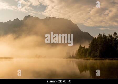 Herbststimmung an einem kleinen See in den bayerischen Alpen, im Hintergrund das Karwendelgebirge, eine kleine Halbinsel im Nebel und die Spiegelung im Wasser. Die neblige Atmosphäre mit den zerklüfteten Bergen hat eine fast mystische Wirkung. Stockfoto