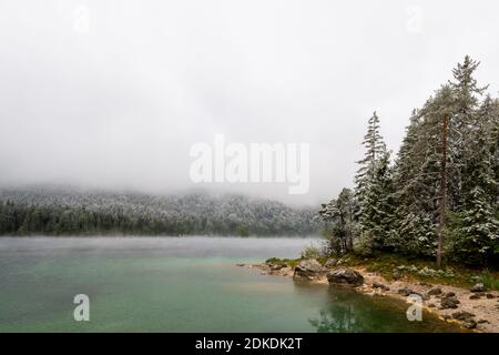 Nebel, Wolken und frischer Schnee am Ufer des schönen Eibsee unterhalb der Zugspitze in den bayerischen Alpen. Stockfoto