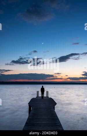 Ein einzelner Mann im Hoodie steht bei Sonnenuntergang mit einem Halbmond auf einem hölzernen Steg am Ufer des Ammersee. Stockfoto