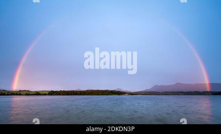 Ein großer Regenbogen zieht sich in satten Farben über den Riegsee, im Hintergrund das Estergebirge mit dem Herzogstand und Jochberg. Stockfoto
