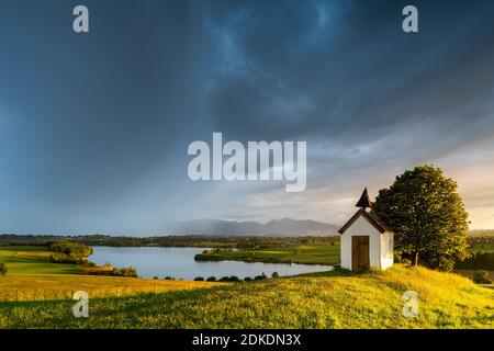 Sturm bei Sonnenuntergang am Riegsee, im Hintergrund die deutschen Alpen des Karwendels, des Estergebirges und der Ammergauer Alpen - mit dunklen Sturmwolken und Regenschleiern, sowie einem schönen Sonnenstern im Tiefland. Im Vordergrund die kleine Sakristan-Kapelle mit ihrem krummen Holzturm und einer saftig grünen Wiese. Stockfoto