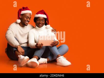 Glückliches schwarzes Paar in Santa Hüte mit digitalen Tablet auf Orangefarbener Hintergrund Stockfoto