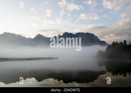 Herbststimmung an einem kleinen See in den bayerischen Alpen, im Hintergrund das Karwendelgebirge, eine kleine Halbinsel im Nebel und die Spiegelung im Wasser. Die neblige Atmosphäre mit den zerklüfteten Bergen hat eine fast mystische Wirkung. Stockfoto