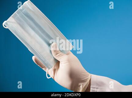 Gesichtsmaske in Arzthand mit OP-Handschuh und blauem Hintergrund. Stockfoto