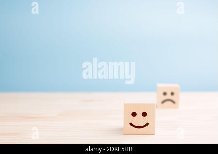 Holzwürfel mit lächelnden Emoticons und traurigen auf Holztisch, blauer Hintergrund. Raum für Text. Emotionales Zustandskonzept Stockfoto