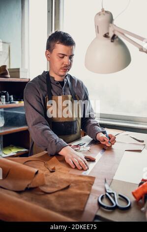 Ein junger Lehrling nimmt Messungen auf Leder in der Werkstatt auf dem Tisch mit einem Lineal. Stockfoto