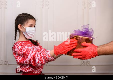 Kindheit und Menschen, das Konzept von Weihnachten während der Epidemie. Mädchen in einer Maske und Handschuhe erhält ein Geschenk. Stockfoto