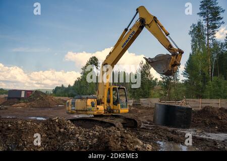 Der Bagger trägt einen Betonring, um auf einer Baustelle einen Kanalisationsbrunnen in eine Rohrleitung zu installieren. Stockfoto