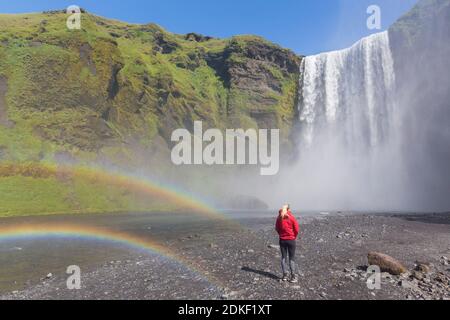 Junge Frau / Tourist vor Skogafoss, 63 m hoher Wasserfall am Fluss Skógá mit doppeltem Regenbogen im Sommer, Skógar, Island Stockfoto