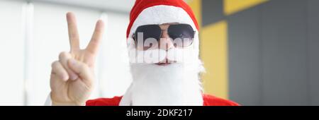 Mann im weihnachtsmann Kostüm mit weißem Bart und Sonnenbrille hält seine Finger im Zeichen des Sieges. Stockfoto