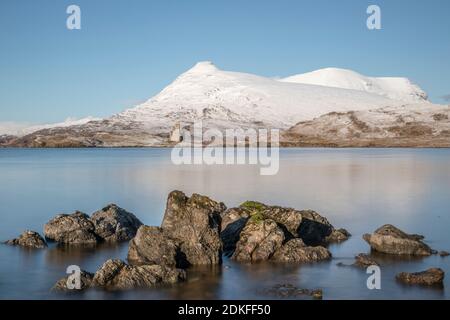 Blick über Steine am Ufer des winterlichen Loch Assynt, Schottland, Ruine von Ardvreck Castle mit schneebedeckten Bergen im Hintergrund Stockfoto