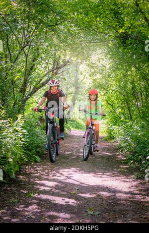 Italien, Venetien, Belluno, Agordino, lächelnde Mutter und Tochter fahren mit dem Fahrrad eine Straße entlang zwischen den Bäumen Stockfoto