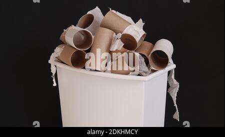 Gebrauchte und leere Toilettenpapierrollen aus Pappe werden in einem kleinen Abfalleimer, isoliert vor schwarzem Hintergrund, aufgestapelt dargestellt. Stockfoto