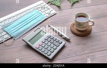 Holztisch mit Tastatur, Gesichtsmaske, schwarzem Kaffee, Stift, Rechner und grünen Pflanzen. Stockfoto