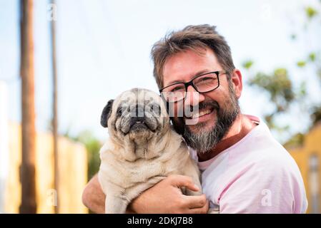 Menschen und Hunde lieben Konzept - Lustiges und glückliches Porträt Von jungen schönen erwachsenen Mann und besten Freund für immer zusammen Hund Mops sowohl Blick auf die Kamera - im Freien Himmel und parkhintergrund Stockfoto