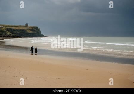 Mann und Frau gehen entlang Sandstrand Nordirland, Castlerock Strand, mit grauem Himmel über dem Atlantik und Mussendun Tempel auf Downhill Klippe. Stockfoto