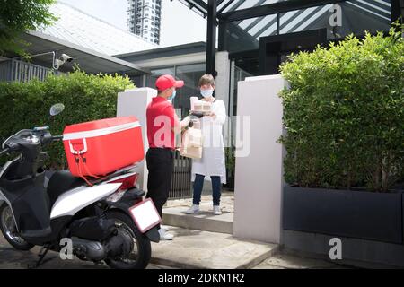 Asiatische Lieferung Mann trägt Gesichtsmaske und Handschuhe in rot Einheitliche Lieferung Beutel mit Lebensmitteln und Getränken an Empfänger während COVID-19-Ausbruch Stockfoto