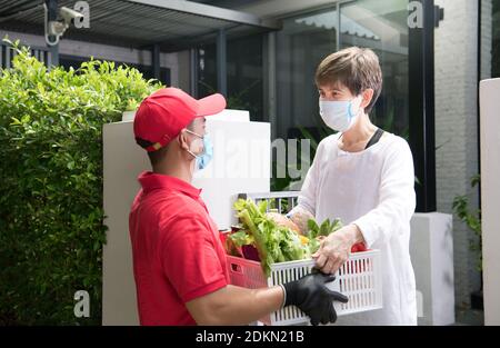Asiatische Lieferung Mann trägt Gesichtsmaske und Handschuhe in roter Uniform Lieferung Lebensmittel-Box mit Lebensmitteln, Obst, Gemüse und Getränke an den Empfänger während COVID Stockfoto