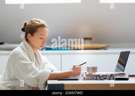 Junge Unternehmerin trägt kuscheligen warmen Bademantel, der in der Weihnachtszeit im Winter während der 2020 Corona-Virus-Pandemie fernab von zu Hause arbeitet. Arbeiten von Stockfoto