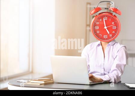 Junge Geschäftsfrau mit Wecker statt Kopf am Tisch im Büro. Zeitmanagement-Konzept Stockfoto