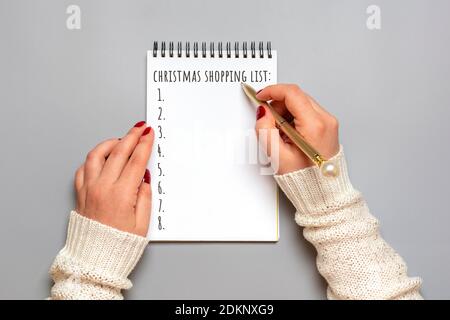 Weibliche Hand hält Stift, schreiben Weihnachtseinkaufsliste, Geschenkideen auf weißem Notizblock auf grauem Hintergrund Draufsicht Flat Lay Weihnachtskarte Frau macht Chr. Stockfoto