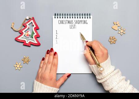 Weibliche Hand hält Stift, schreiben Weihnachtseinkaufsliste, Geschenkideen auf weißem Notizblock auf grauem Hintergrund Draufsicht Flat Lay Weihnachtskarte Frau macht Chr. Stockfoto