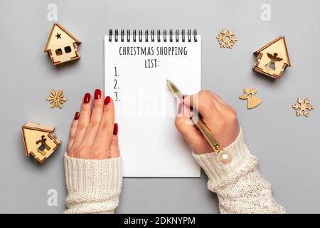 Weibliche Hand hält Stift, schreiben Weihnachtseinkaufsliste, Geschenkideen auf weißem Notizblock auf grauem Hintergrund Draufsicht Flat Lay Weihnachtskarte Frau macht Chr. Stockfoto